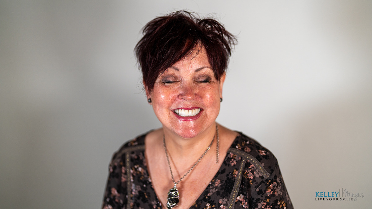 Woman with short dark hair and floral blouse smiles with eyes closed against a light background, radiating confidence often seen after Full Mouth Rehabilitation in Bend Oregon. She wears a pendant necklace, and there is a watermark in the bottom right corner.