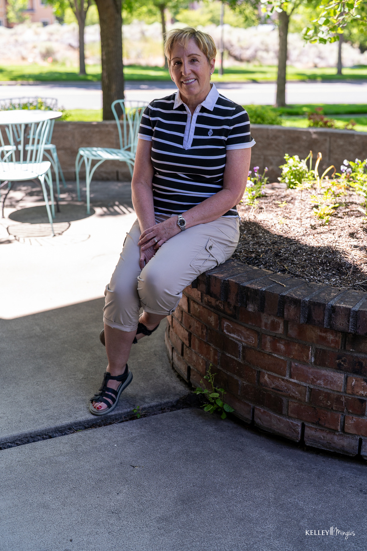 A person wearing a striped polo shirt and khaki pants sits on a brick planter in an outdoor patio area with metal chairs and tables nearby, perhaps taking a break after their recent teeth cleaning appointment or contemplating options in cosmetic dentistry.