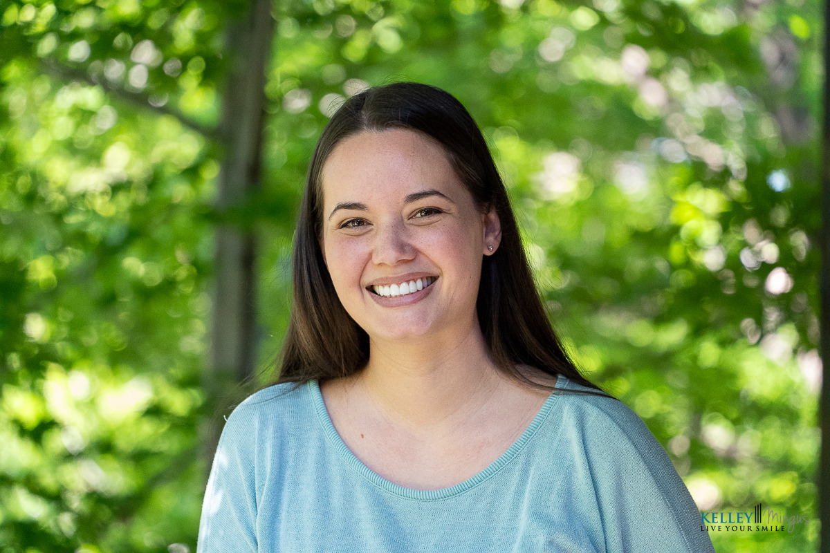 A woman with long brown hair and a light blue top smiles outdoors, with green trees blurred in the background—radiating the confidence that comes from expert care by Kelley Mingus, a renowned TMJ specialist.