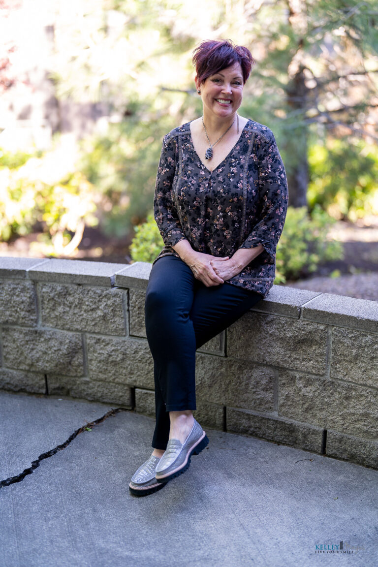 A woman with short hair, wearing a patterned blouse and black pants, sits on a stone ledge outdoors, smiling at the camera—she exudes calm confidence, much like someone who has just received expert care from Kelley Mingus TMJ specialist.