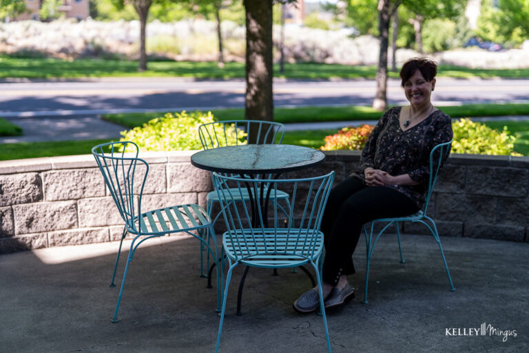 A woman sits at a round metal patio table with three empty chairs outside on a sunny day, with trees and greenery in the background, enjoying a peaceful moment after her visit to Kelley Mingus TMJ specialist.