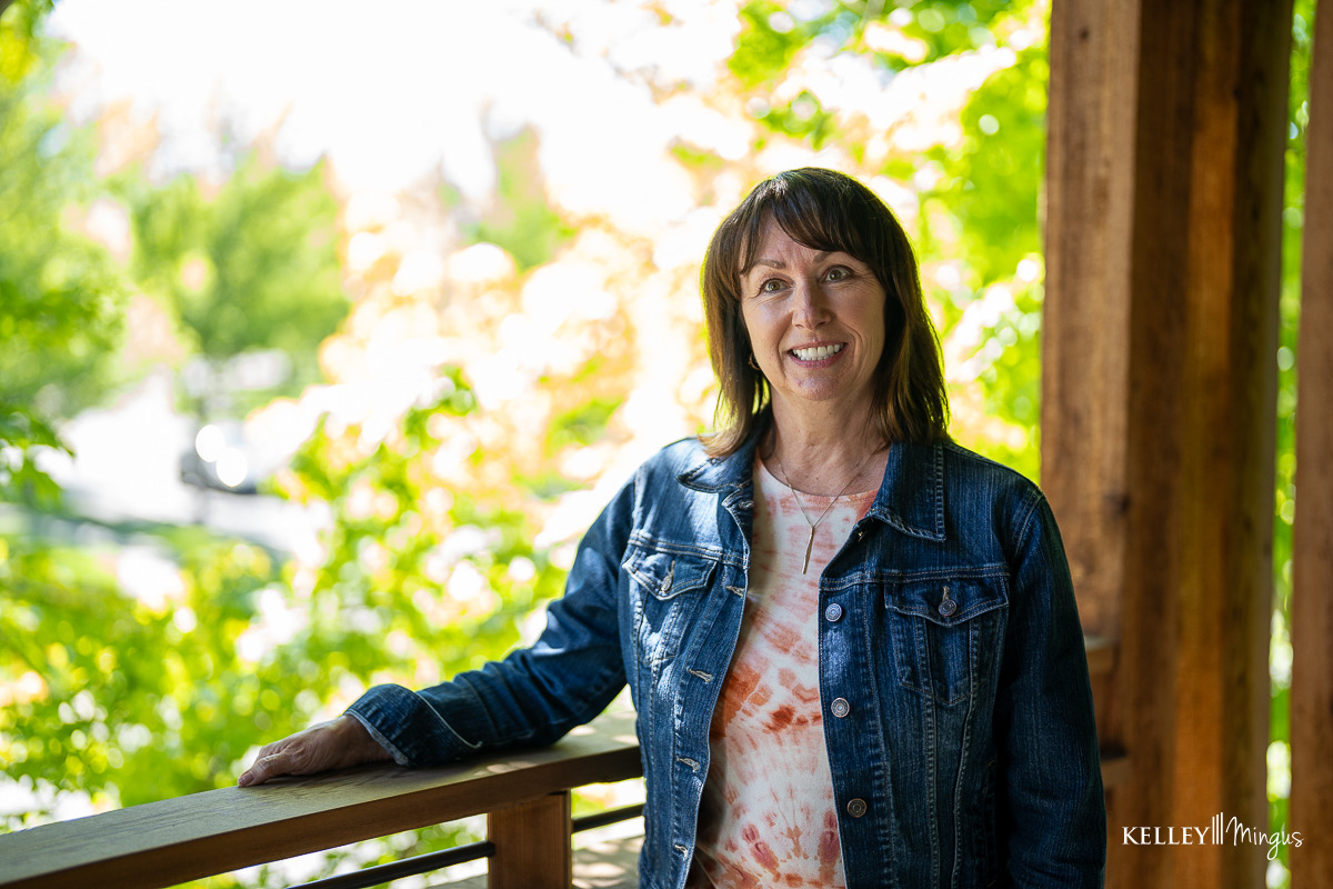 A smiling woman wearing a denim jacket stands on a porch with green foliage in the background, a person who might visit a clinic for affordable cosmetic dentistry in Bend.