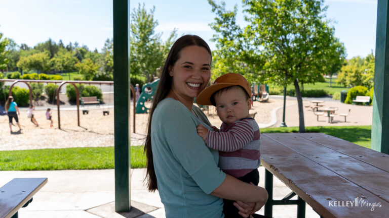 A woman with a bright smile, perhaps benefiting from affordable cosmetic dentistry in Bend, holds a baby in a cap at a park picnic table.