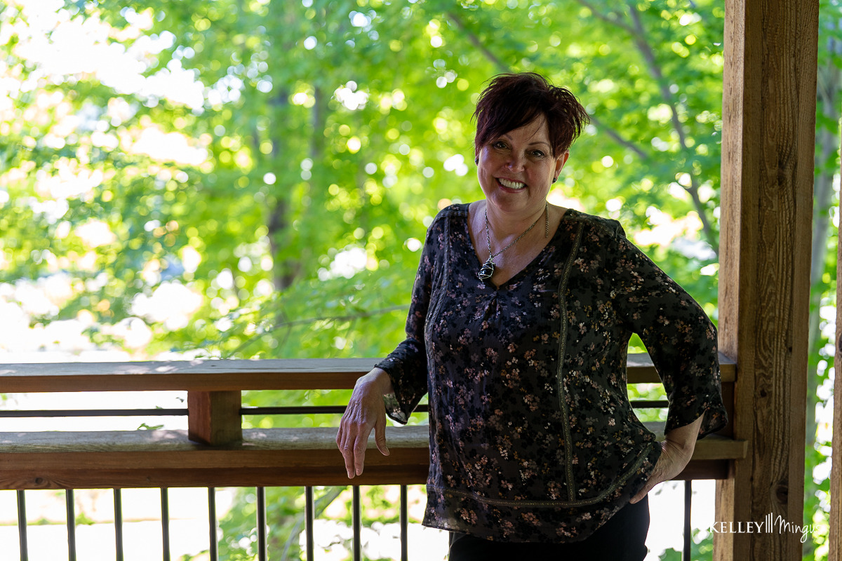 A woman with short dark hair, wearing a patterned blouse, stands and smiles on a wooden deck with trees behind her, looking relaxed like a Kelley Mingus TMJ specialist.