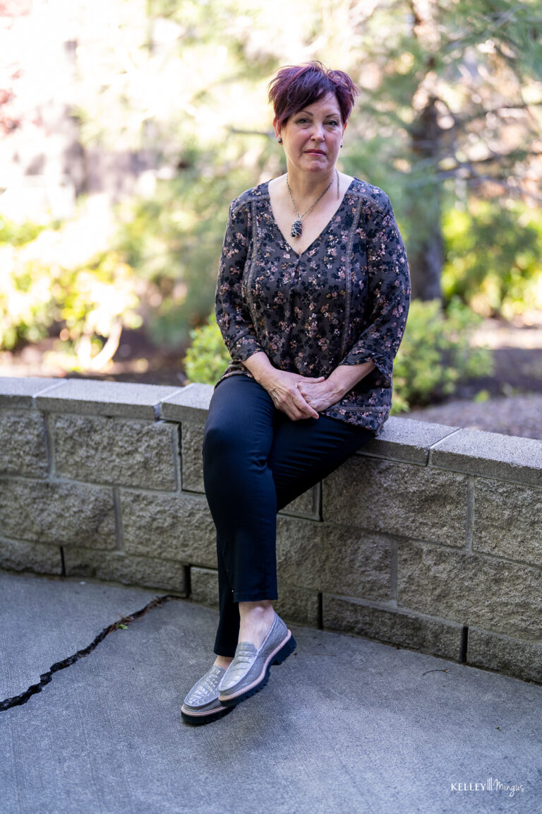 A woman with short dark hair and a bright smile indicative of professional teeth cleaning sits on a stone wall, wearing a patterned long-sleeved top, black pants, and metallic slip-on shoes.
