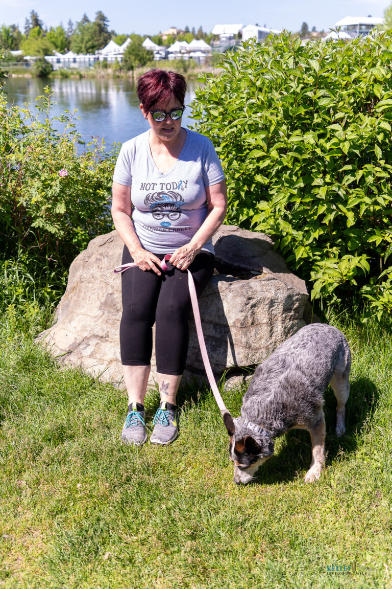A woman sits on a rock by a lake holding a leash attached to a blue heeler sniffing the grass, resting before researching CPAP alternatives for sleep apnea.