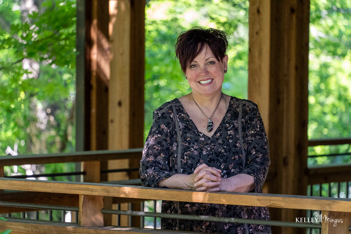 A woman with short, dark hair and a bright smile for cosmetic dentistry wears a floral print top and a pendant necklace while leaning against a wooden railing.