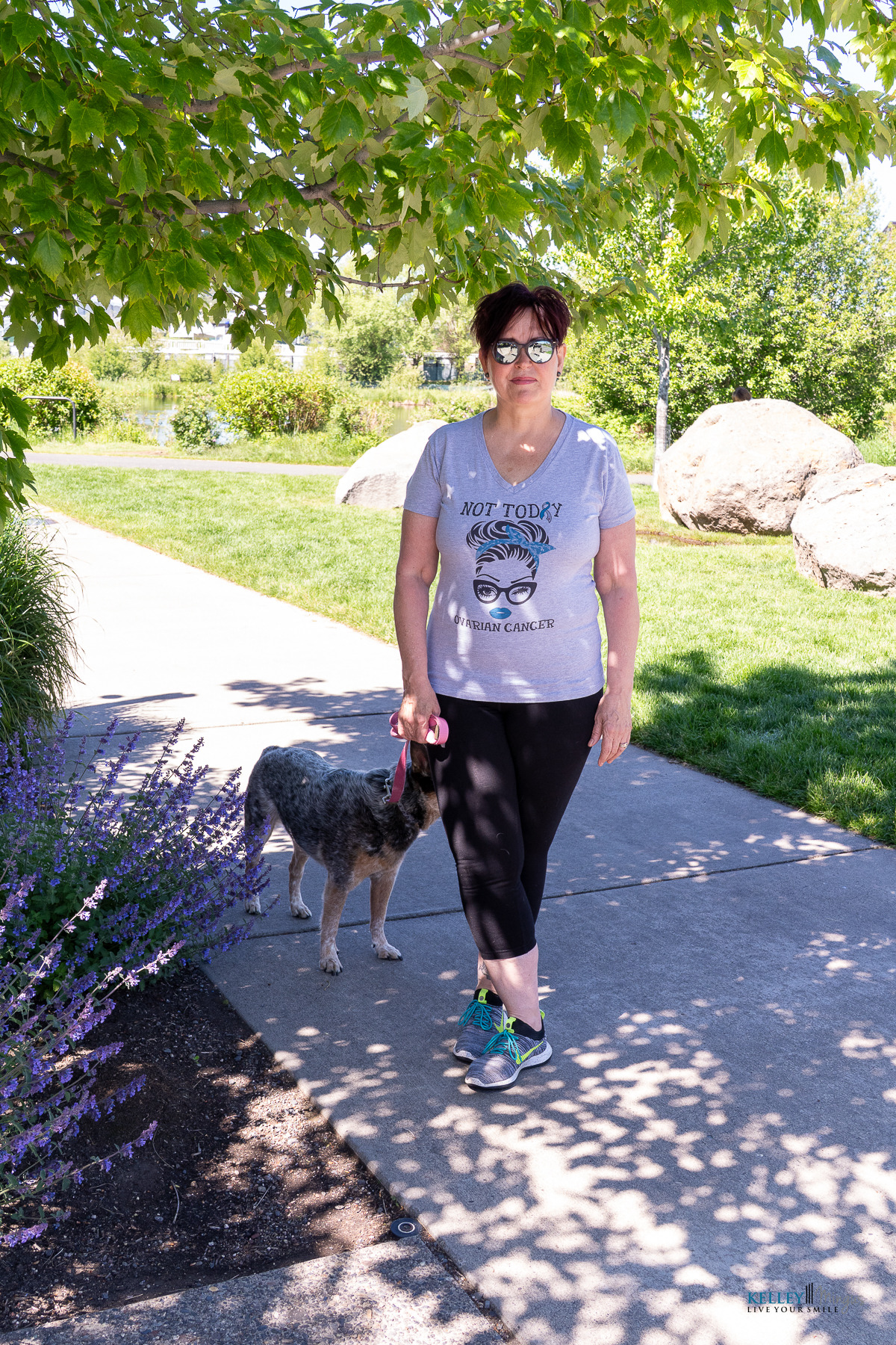 A woman wearing sunglasses and an ovarian cancer awareness t-shirt stands on a park path holding the leash of a mottled dog, perhaps seeking relief from conditions like CPAP alternatives for sleep apnea.