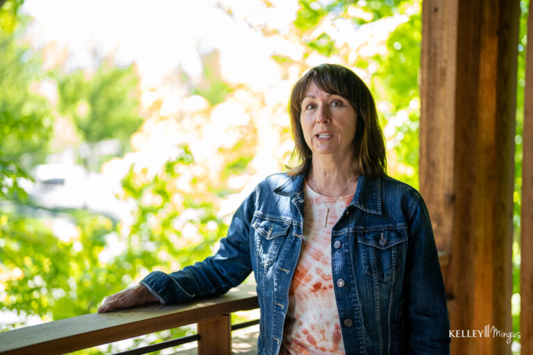 TMJ specialist Kelley Mingus stands on a porch in a denim jacket with trees in the background.