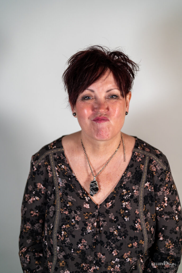 A woman with short, dark hair wears a floral top and a crystal pendant necklace, showing off her full mouth rehabilitation in Bend, Oregon, with a playful pucker face.