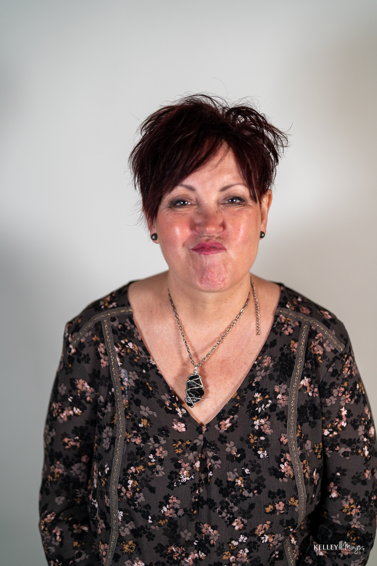 A woman with short, dark hair wears a floral top and a crystal pendant necklace, showing off her full mouth rehabilitation in Bend, Oregon, with a playful pucker face.