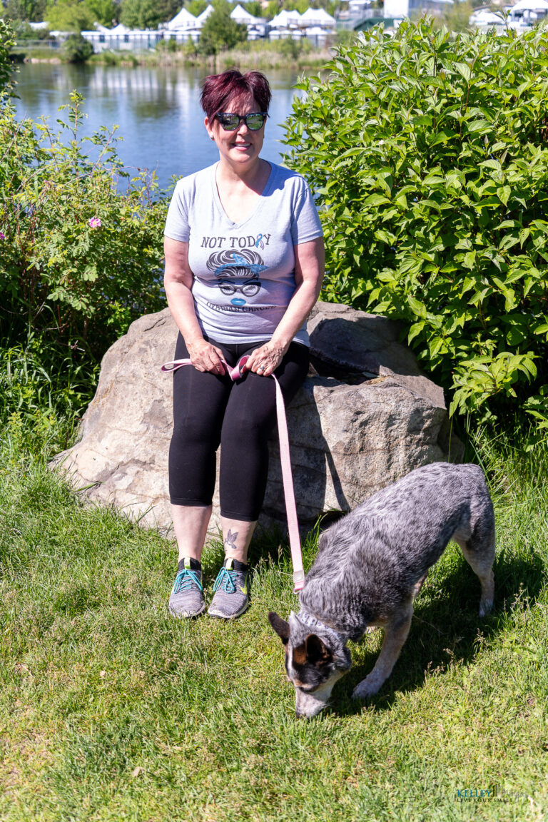A woman sits on a rock by a lake in Bend, Oregon, holding a leash attached to a blue heeler sniffing the grass.