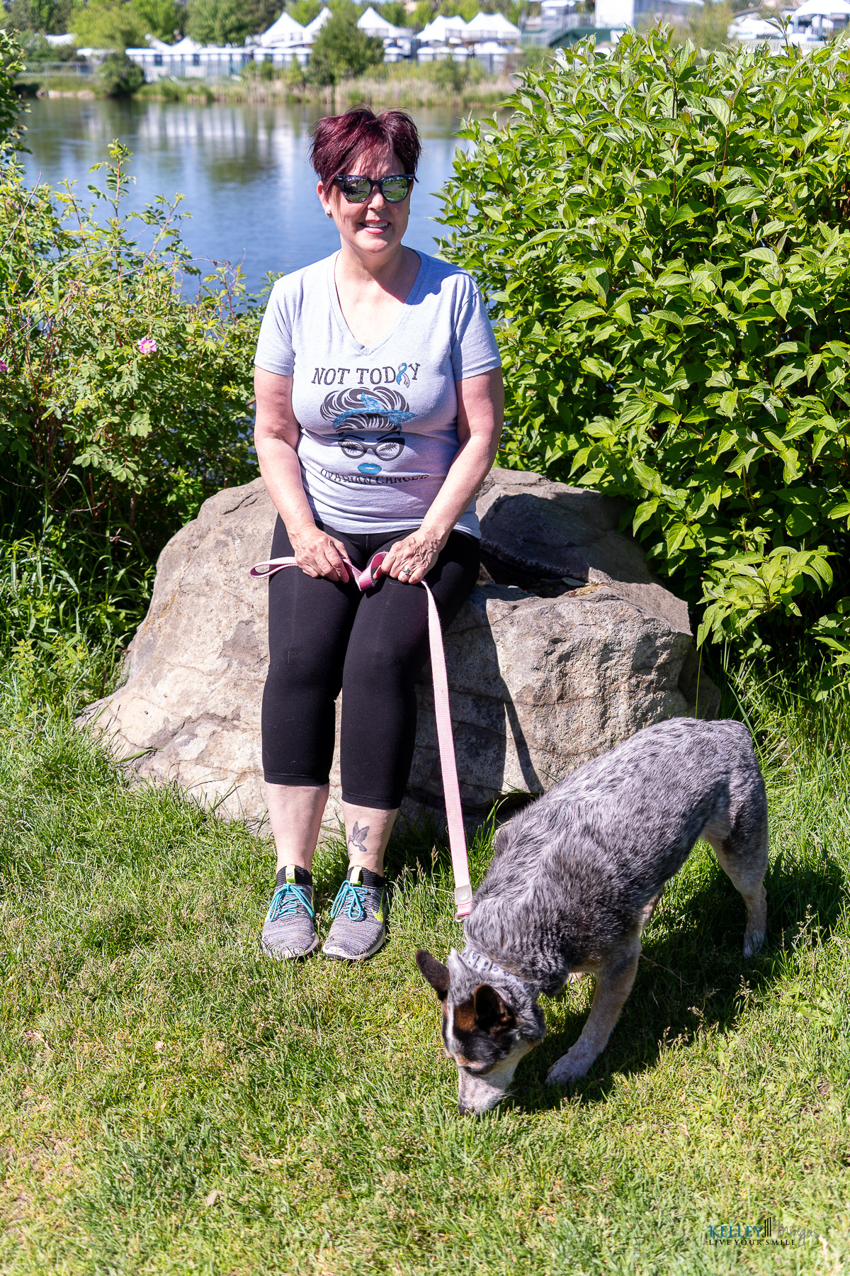 A woman sits on a rock by a lake in Bend, Oregon, holding a leash attached to a blue heeler sniffing the grass.